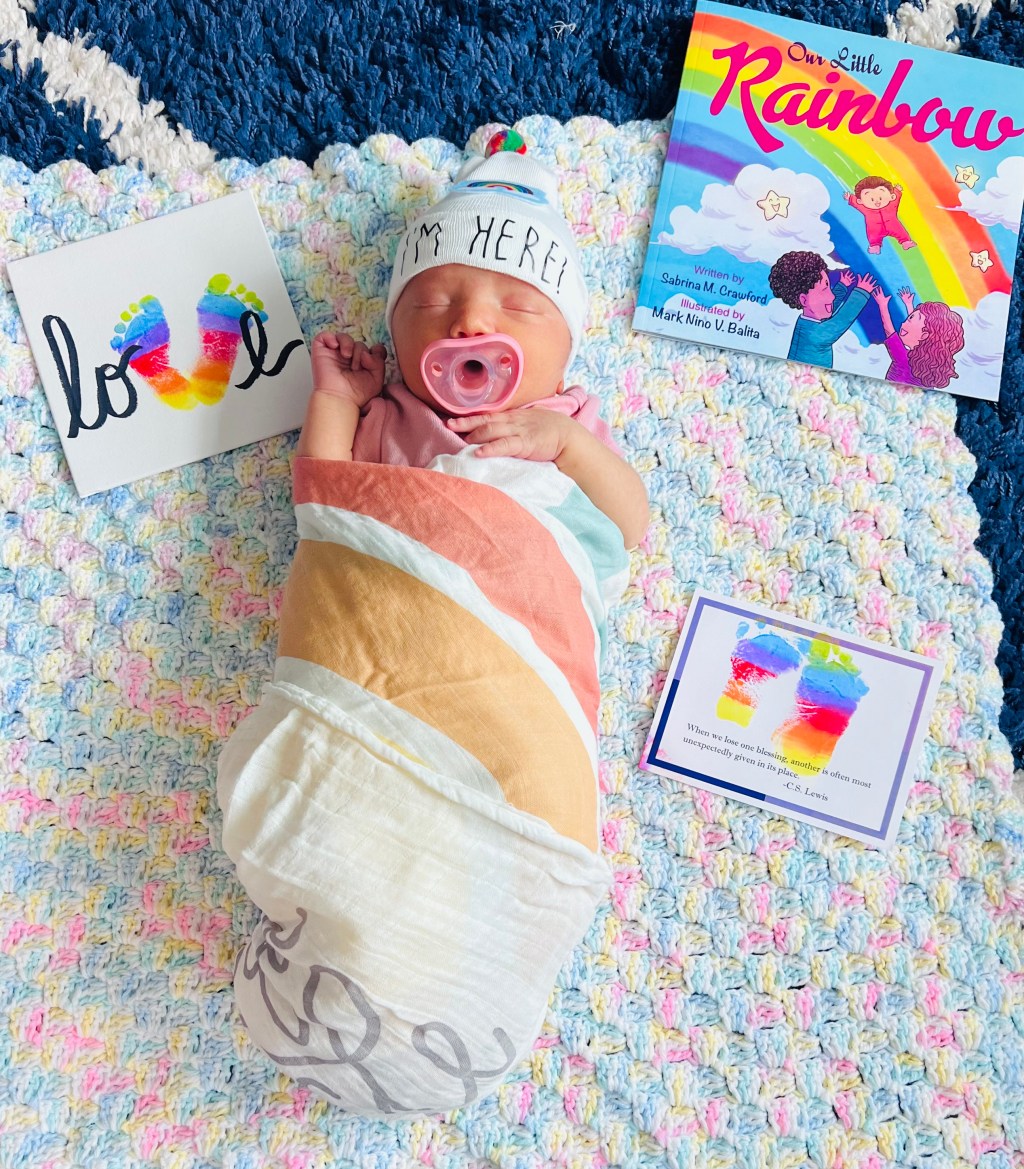 Newborn rainbow baby Samantha lying at home on her crocheted rainbow blanket, with her rainbow-colored footprints and a rainbow-themed book beside her — a symbol of life, healing, and hope after stillbirth.